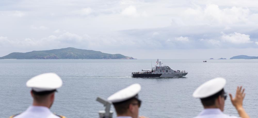 RAN officers and sailors wave as HMAS Arunta sails past a Royal Thai Navy patrol boat.