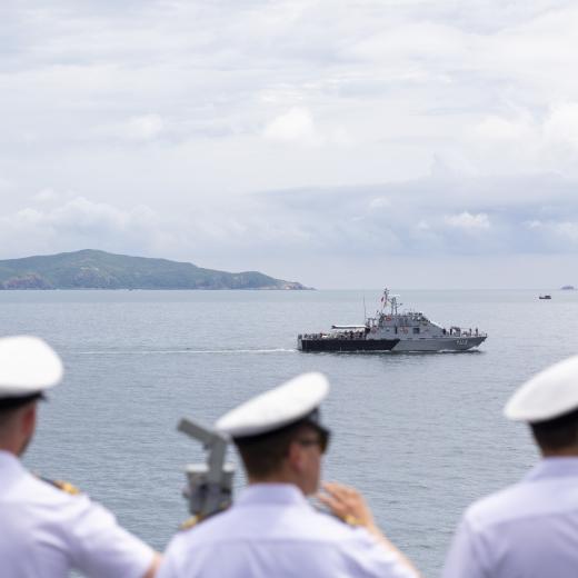 RAN officers and sailors wave as HMAS Arunta sails past a Royal Thai Navy patrol boat.