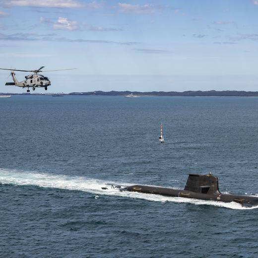 Collins Class submarine heading out to sea from Fleet Base West, shadowed by a  Seahawk 